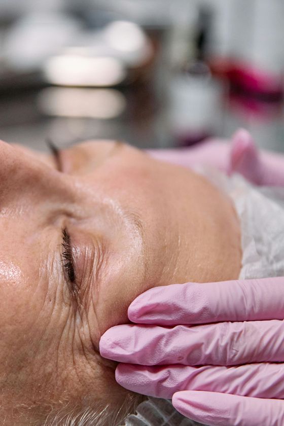 Primer plano del rostro de una persona, tocada con guantes rosas, con una leve sonrisa y un ojo cerrado, en una clínica.