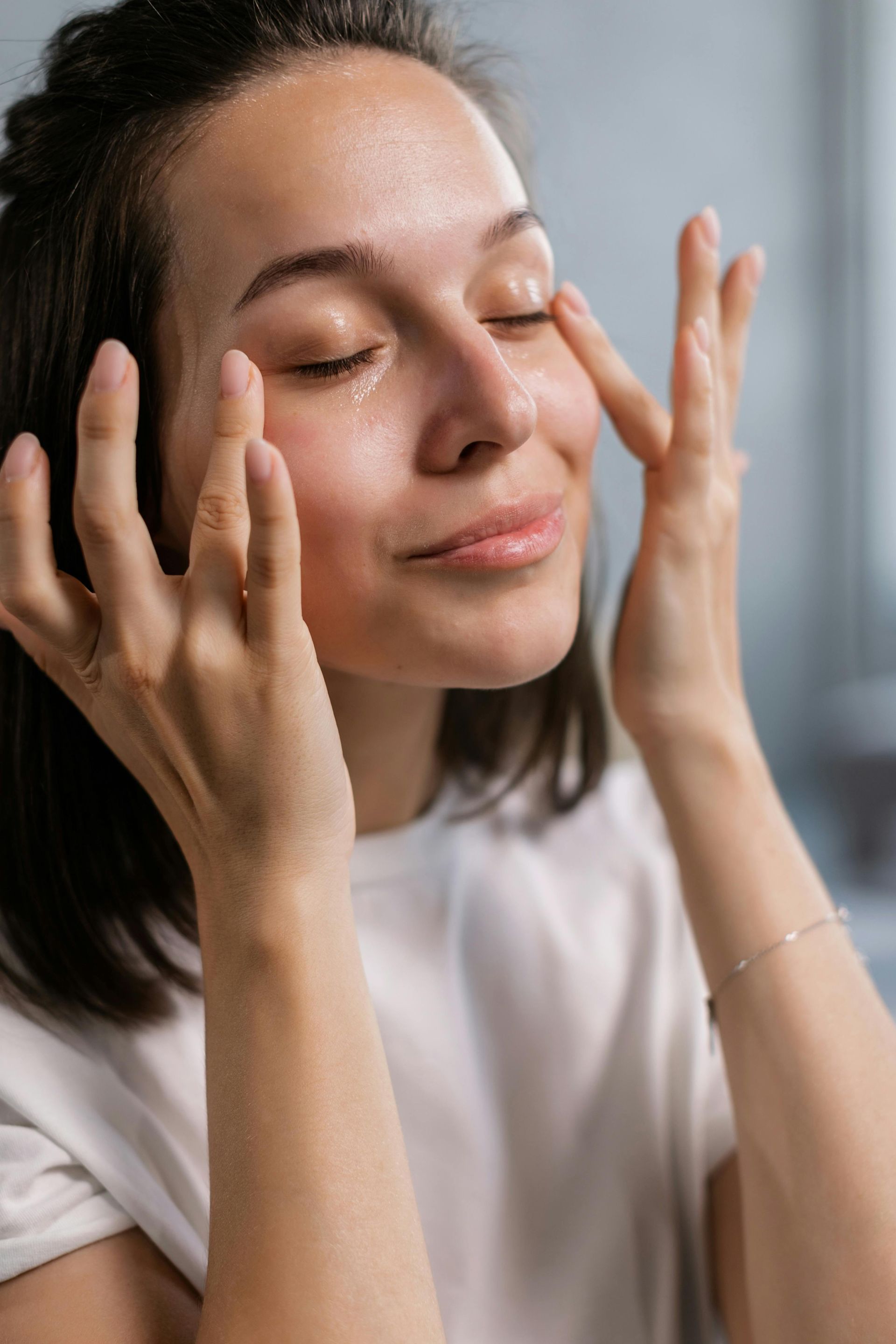 Mujer con los ojos cerrados, aplicándose crema facial con los dedos. Camiseta blanca, piel clara, expresión suave.
