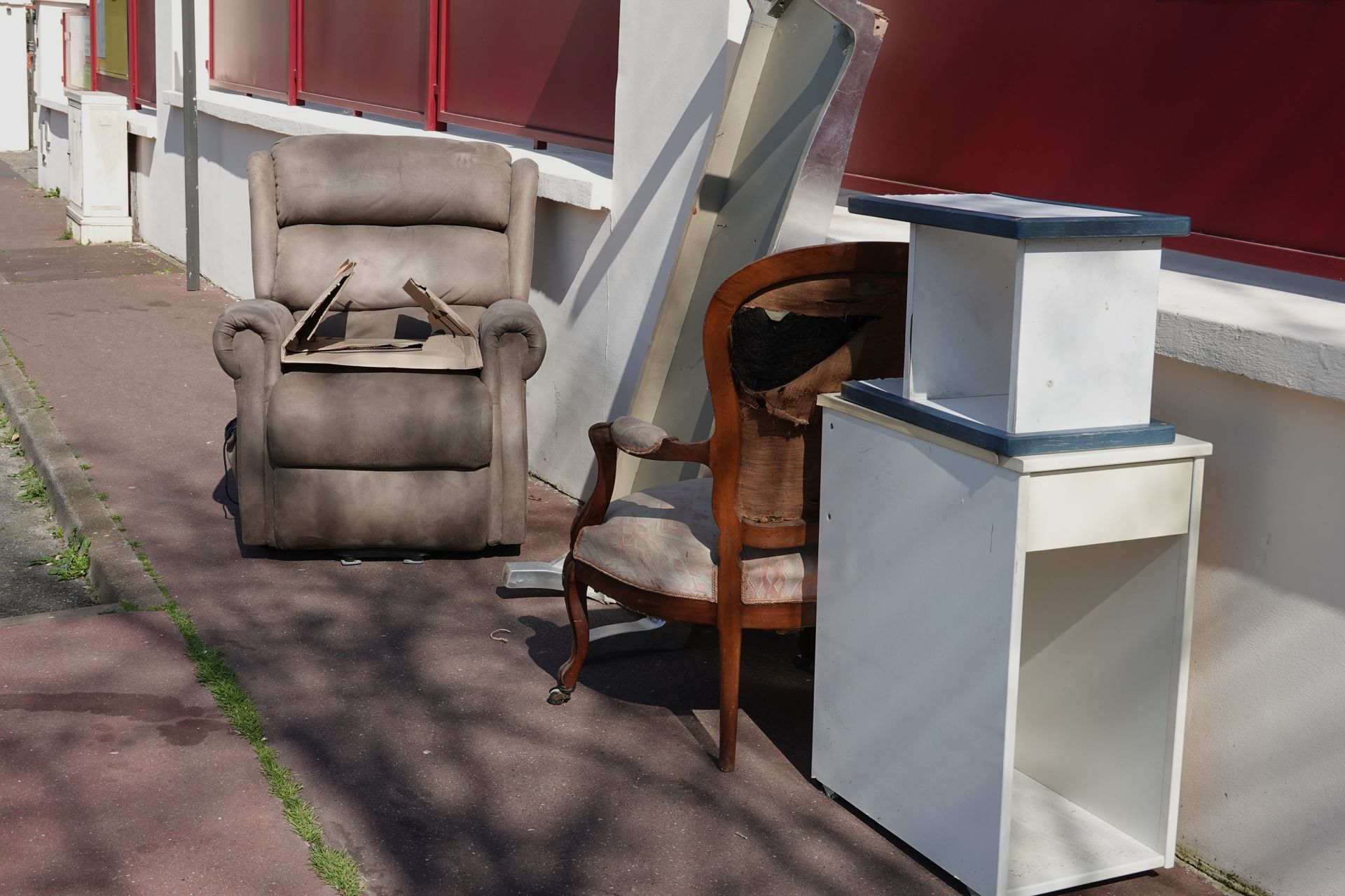 Meubles abandonnés sur un trottoir : fauteuil inclinable, chaise, armoire blanche et planches, à côté d’un bâtiment.