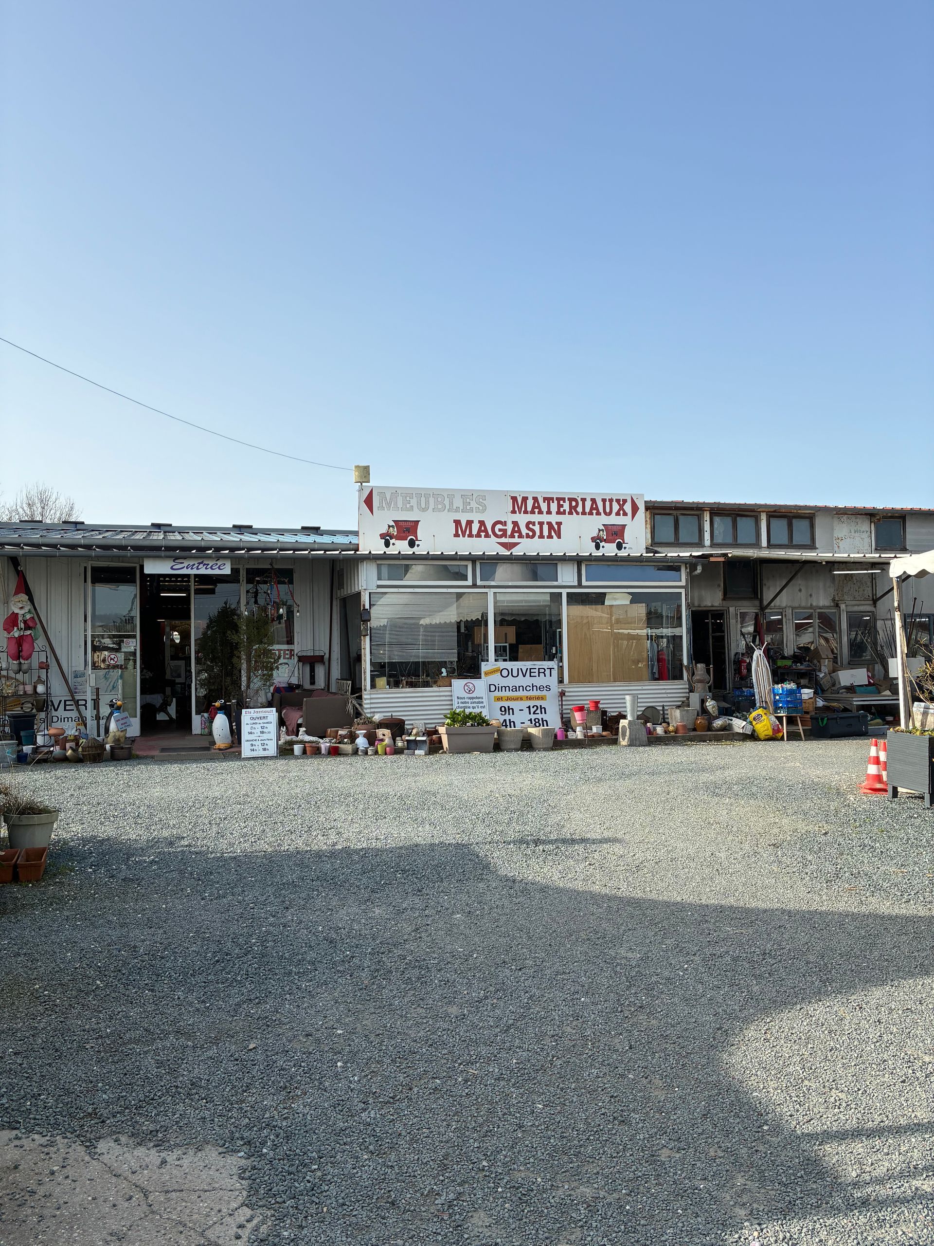 Façade de magasin avec une enseigne en lettres rouges, entourée de gravier, sous un ciel d'un bleu éclatant.