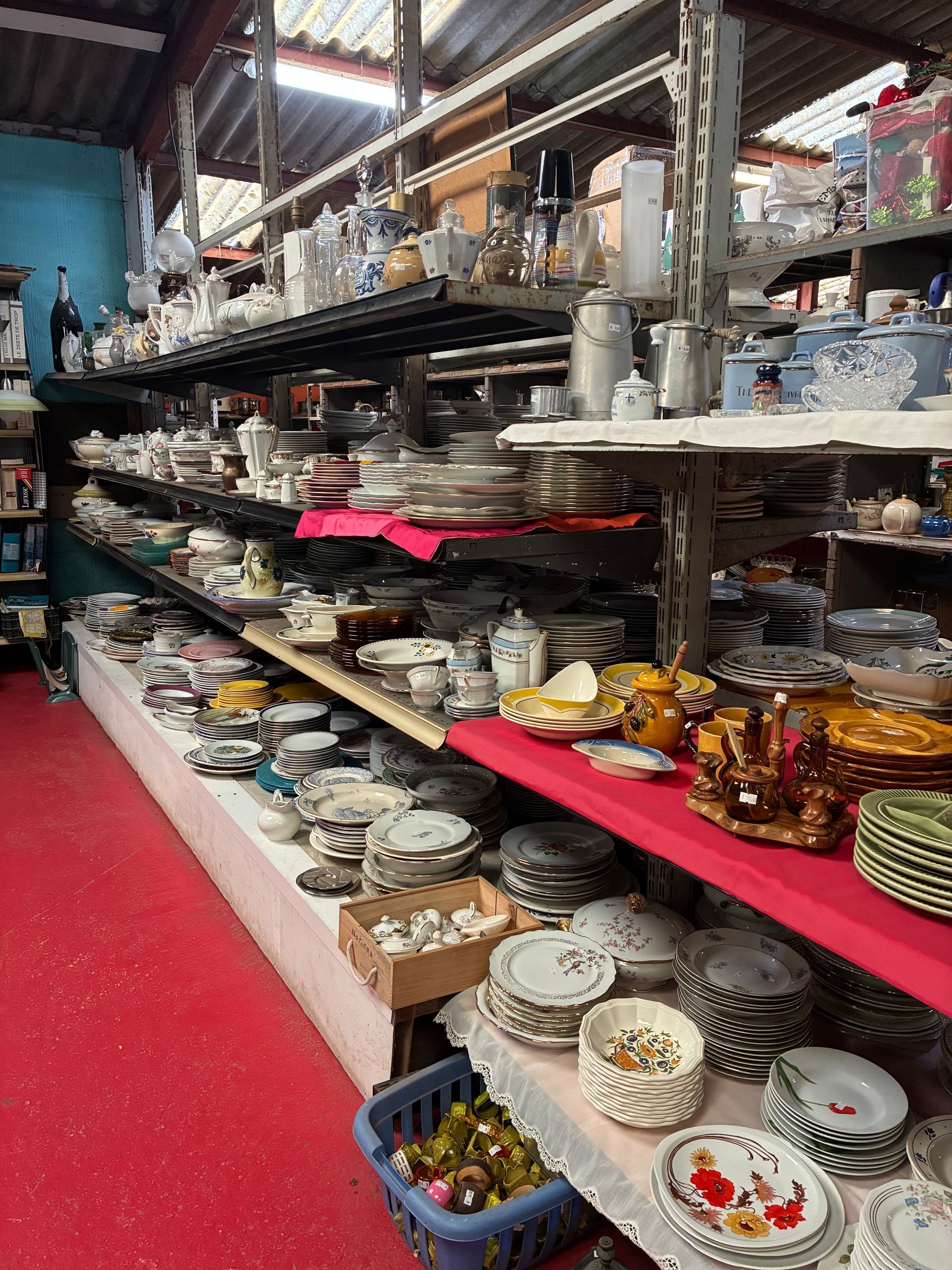 Des étagères regorgent de vaisselle et d'articles de service dans un magasin encombré. Moquette rouge, éclairage zénithal.