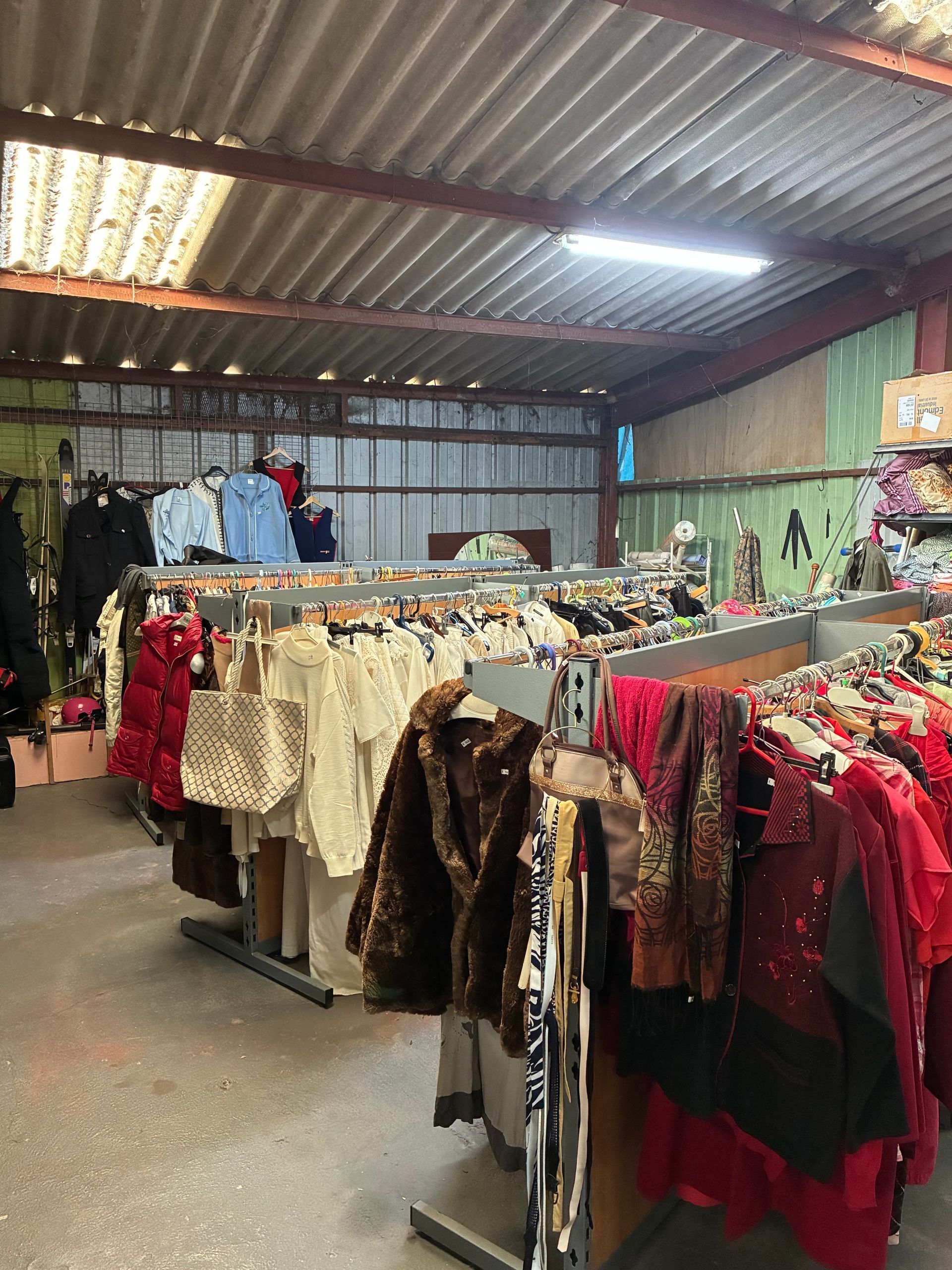 Dans un entrepôt, des portants remplis de vêtements divers sont visibles. La charpente apparente et l'éclairage sont présents.