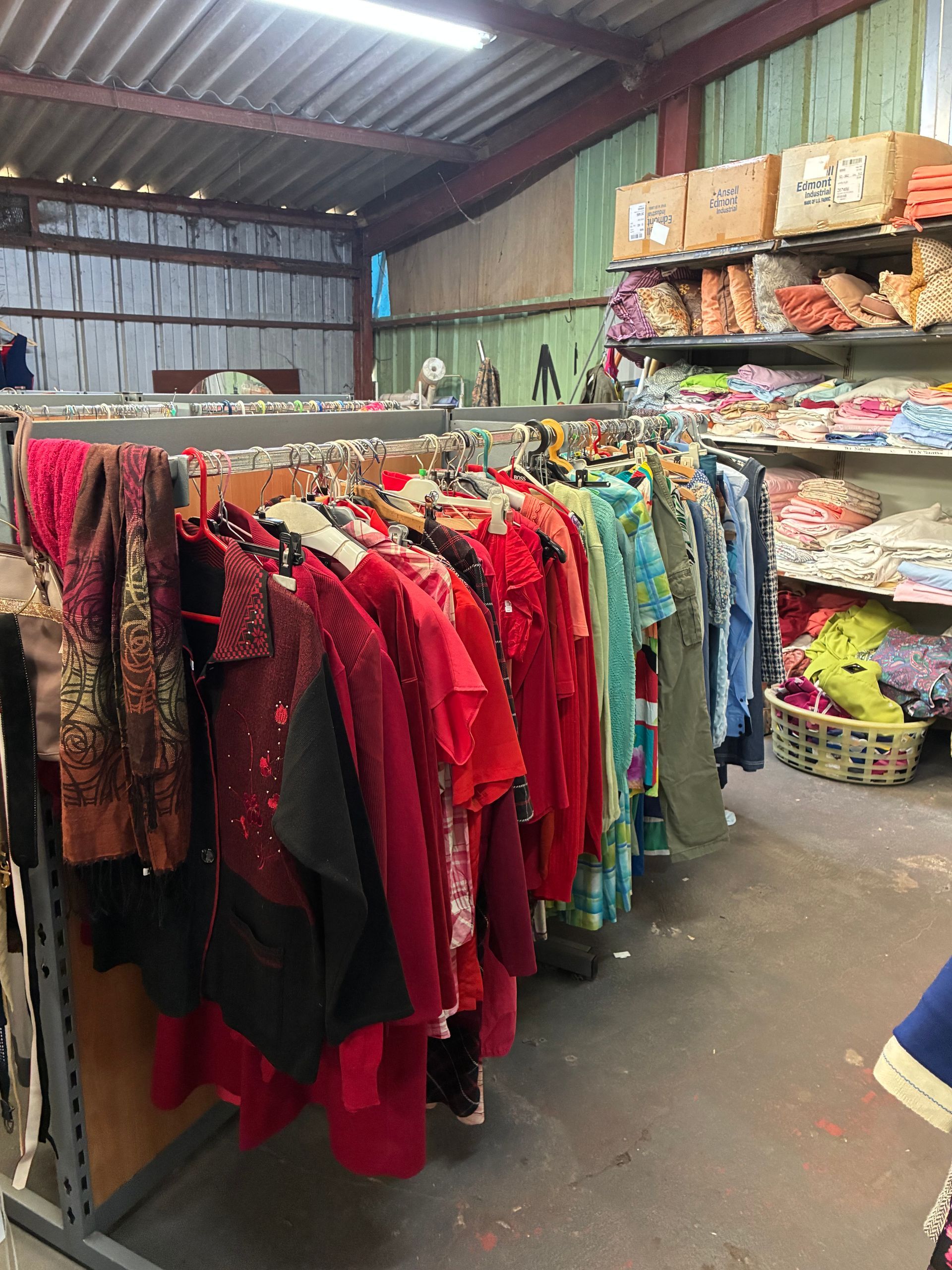 Des portants à vêtements dans un entrepôt lumineux regorgent de vêtements colorés. Des étagères sont chargées de tissus et de marchandises.