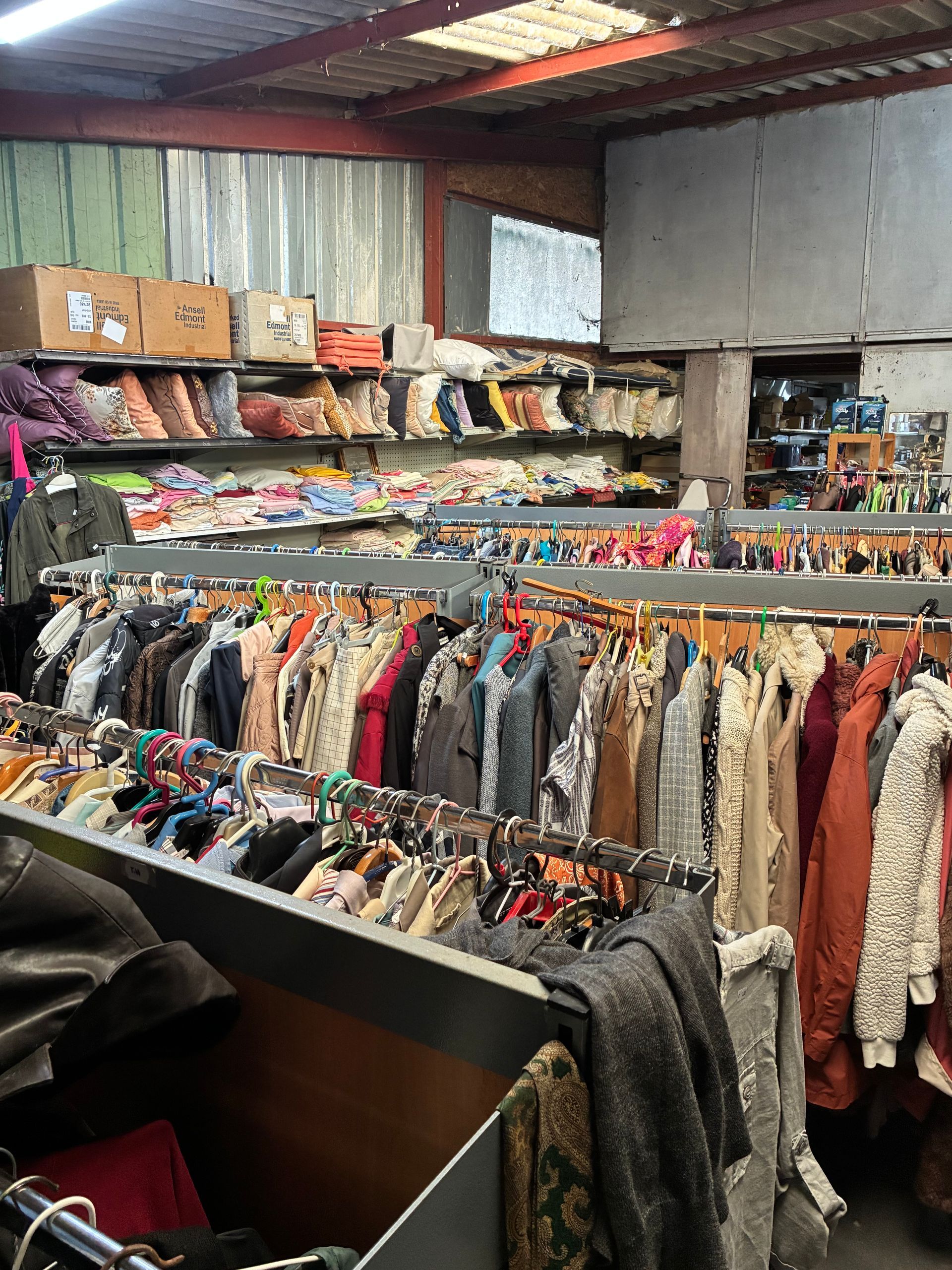 Des portants remplis de vêtements divers dans une friperie. Les étagères du dessus contiennent des chaussures et des boîtes.