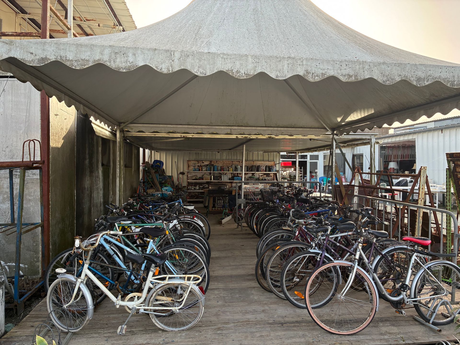 Des vélos sont garés sous une structure ressemblant à une tente dans un espace extérieur, probablement un lieu de stockage ou de location.