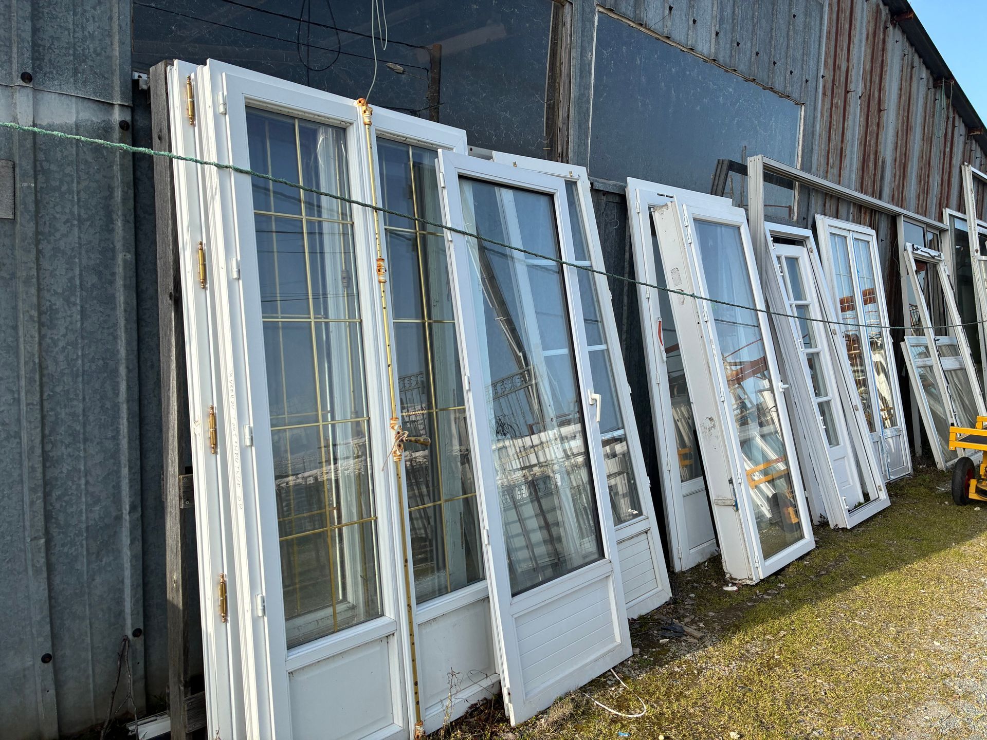 Des portes vitrées à cadre blanc sont appuyées contre un bâtiment gris à l'extérieur, par une journée ensoleillée.