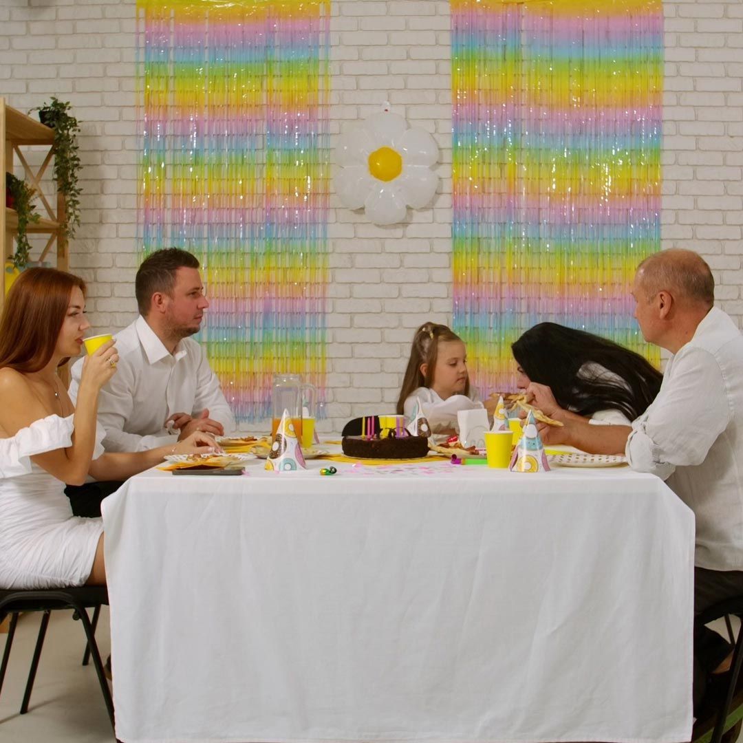 Familia sentada en una mesa con pastel de cumpleaños y bebidas, decorada con serpentinas y globos de colores pastel.