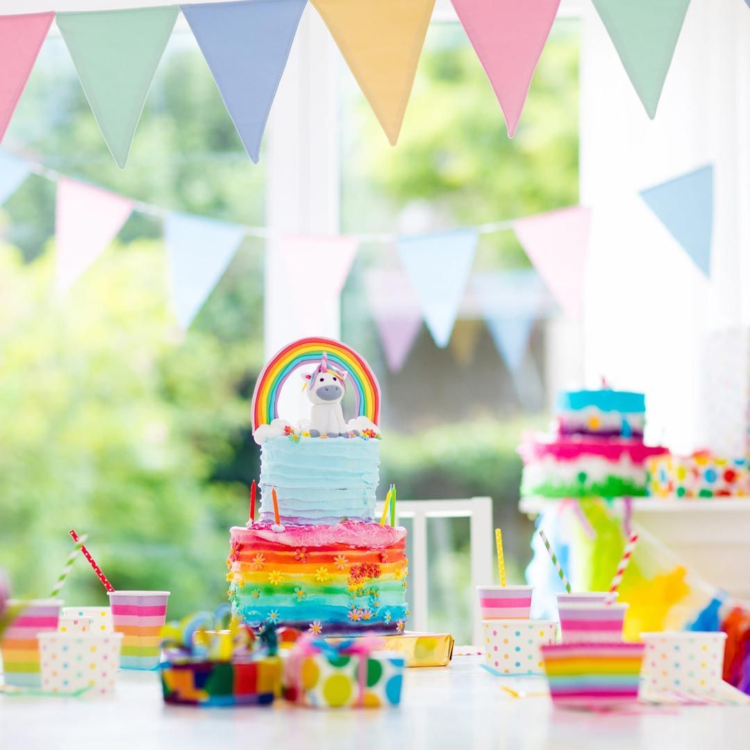 Tarta de cumpleaños de unicornio arcoíris sobre una mesa de fiesta en tonos pastel con guirnaldas coloridas y dulces.