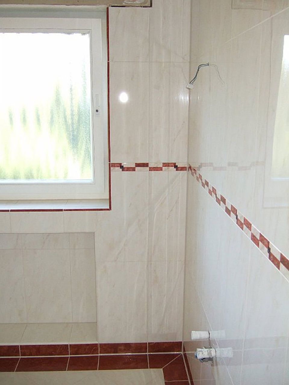 Ein Badezimmer mit weißen Fliesen und roten Zierleisten und einem Fenster Scharwat - Fliesen und Naturstein