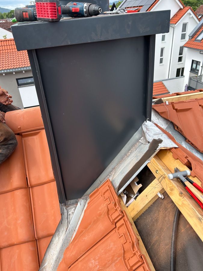 Ein schwarzer Schornstein wird auf einem rot gedeckten Dach montiert; Werkzeuge sind sichtbar.