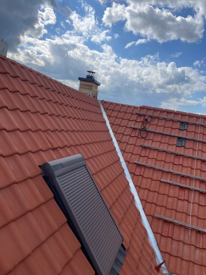 Ein rotes Ziegeldach mit Schornstein, ein geschlossener Dachfensterladen und ein bewölkter blauer Himmel.