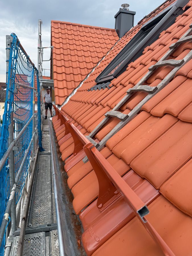 Dach mit orangefarbenen Ziegeln und Dachrinne. Ein Mann steht auf einem Gerüst. Leiter und Oberlicht sind sichtbar.
