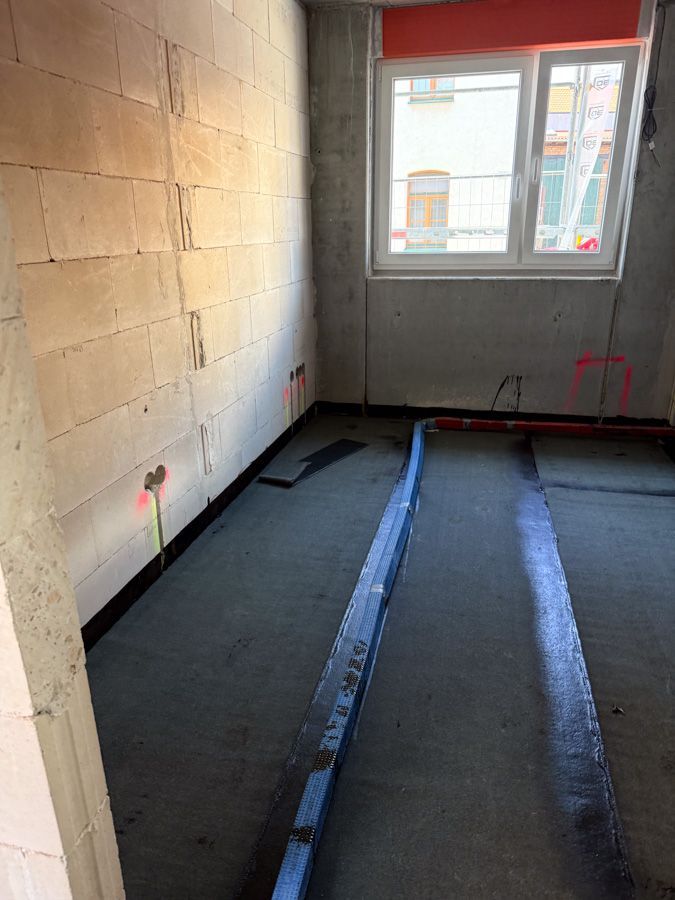 Ein im Bau befindlicher Raum mit Betonblockwänden, einem Fenster und auf dem Boden verlaufenden Wasser- und Elektroleitungen.