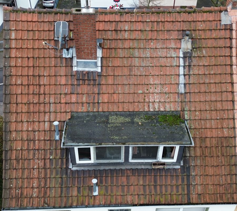 Draufsicht auf ein rotziegelgedecktes Dach mit Dachgaube, Schornstein und Lüftungsrohren. Etwas Moosbewuchs ist sichtbar.
