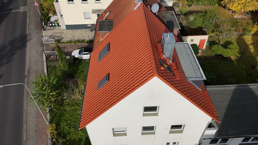 Vogelperspektive auf ein weißes Gebäude mit orangefarbenem Ziegeldach, Schornstein und Oberlichtern. Ein weißes Auto ist in der Nähe geparkt.