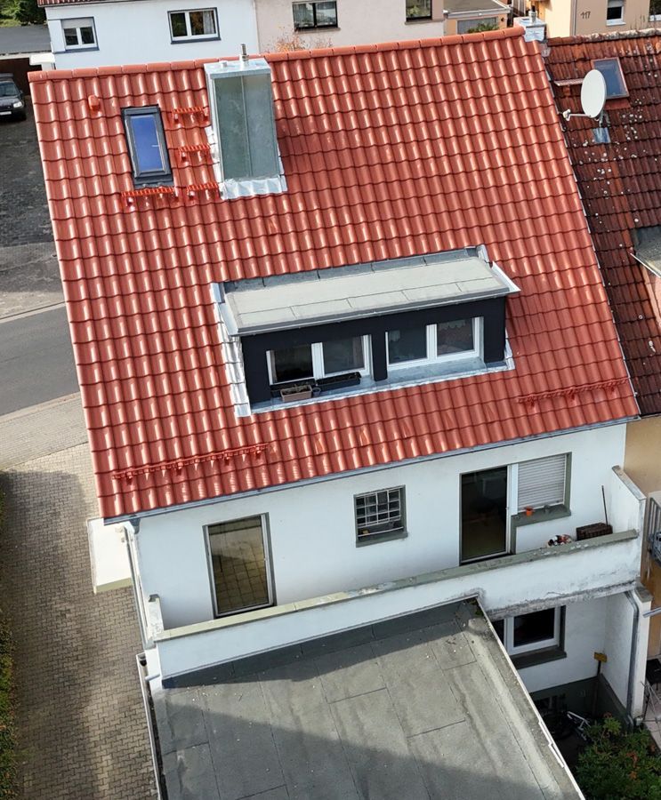 Weißes zweistöckiges Haus mit rotem Ziegeldach, Dachgauben und einem Flachdachanbau.