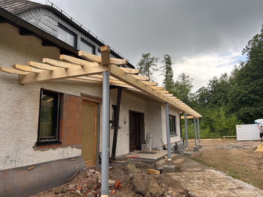 Holzgerüst einer neuen Veranda, die an einem Haus mit grauem Dach und Betonsäulen errichtet wird.