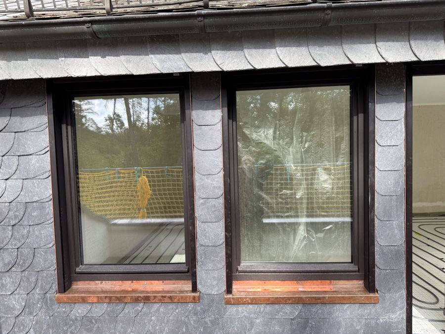 Zwei braun gerahmte Fenster in einem Gebäude mit grauer Schieferverkleidung; kupferfarbene Fensterbänke.