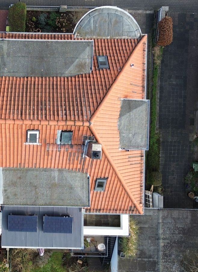Draufsicht auf ein Haus mit orangefarbenem Ziegeldach, Oberlichtern und Solaranlage. Graue Dachabschnitte, ein Ziegelweg und Grünflächen.
