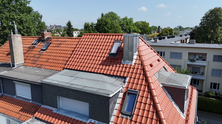 Rotes Ziegeldach mit Schornstein und Oberlichtern, im Hintergrund Gebäude und Bäume an einem sonnigen Tag.