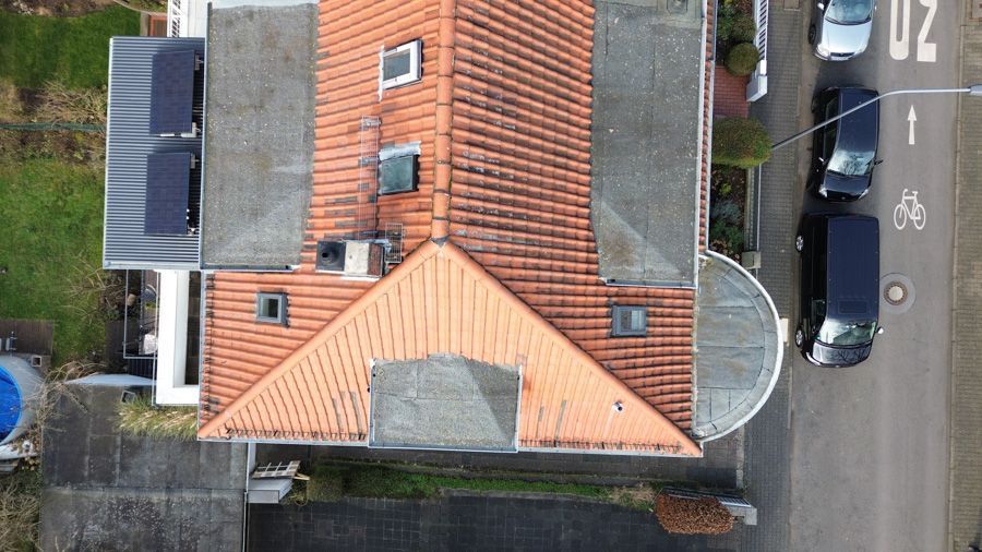 Draufsicht auf ein Haus mit orangefarbenem Ziegeldach, Solarpaneelen und geparkten Autos an einer Straße.