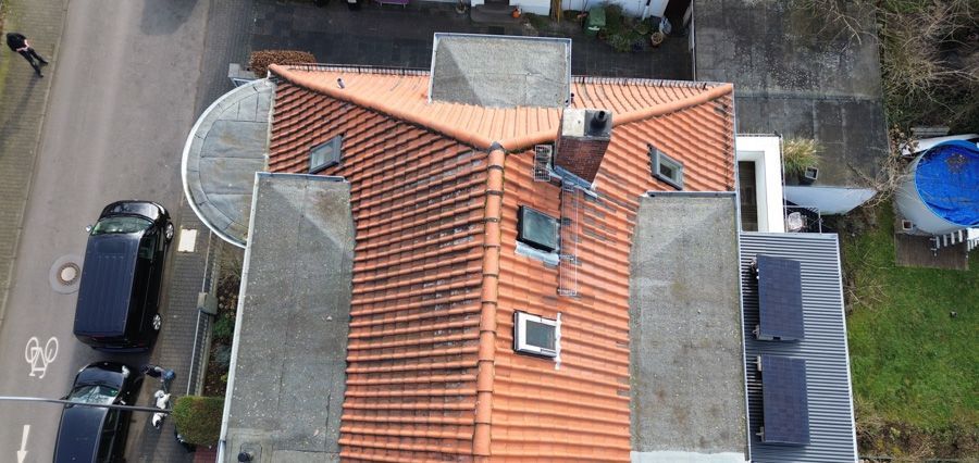 Luftaufnahme eines Hauses mit Ziegeldach, Solarpaneelen und Schornstein. Autos und ein Swimmingpool sind ebenfalls zu sehen.