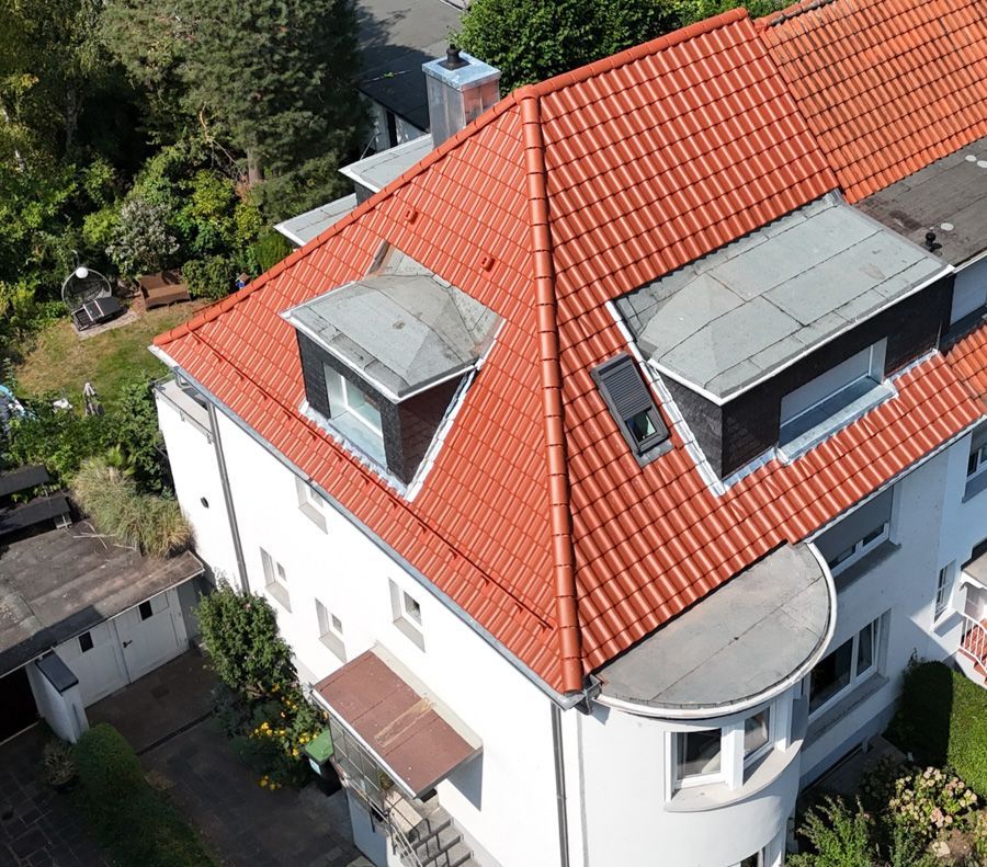 Weißes Gebäude mit rotem Ziegeldach; zwei Dachgauben, umgeben von Bäumen.