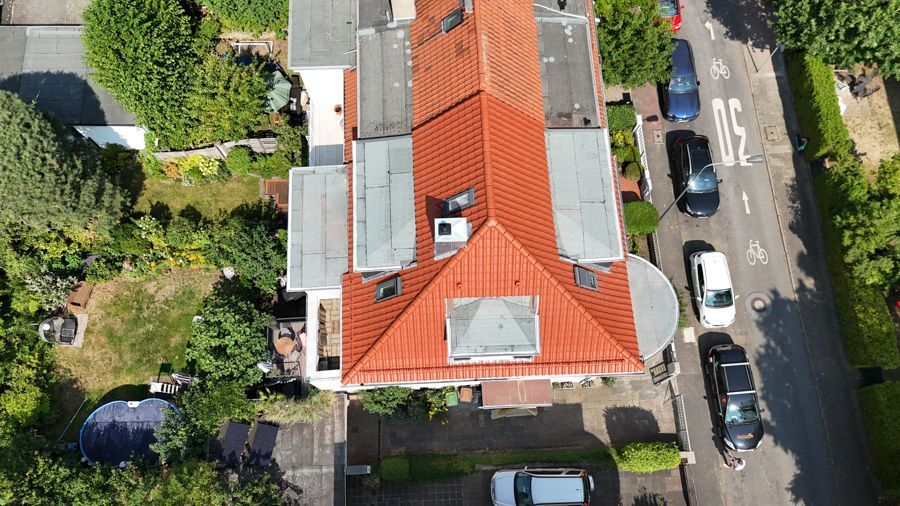 Draufsicht auf ein Haus mit rotem Ziegeldach, umgeben von Bäumen, einer Auffahrt und einer Straße mit geparkten Autos.