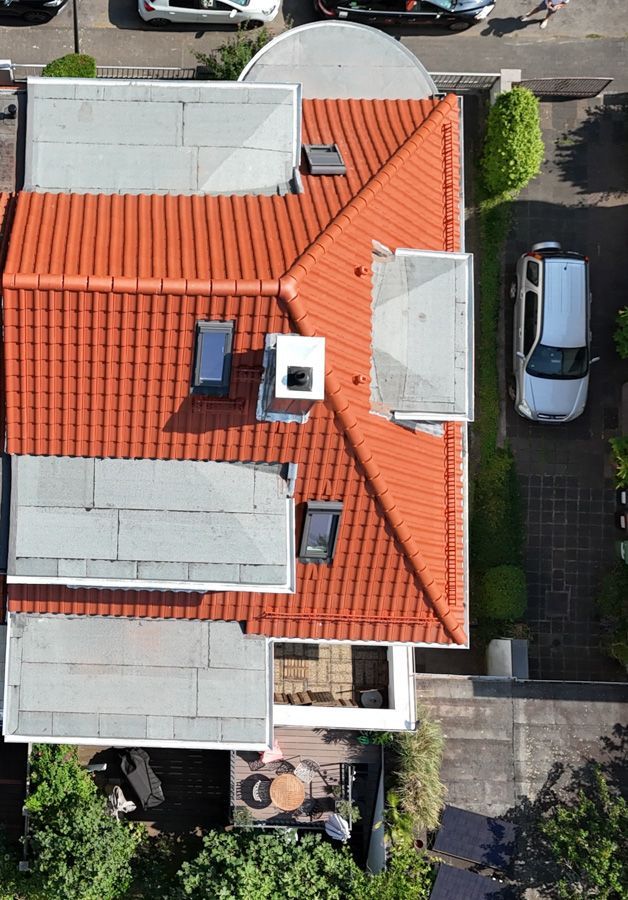 Luftaufnahme eines Hauses mit orangefarbenem Ziegeldach, grauen Flachdächern und einem weißen Schornstein. Autos und Bäume umgeben es.