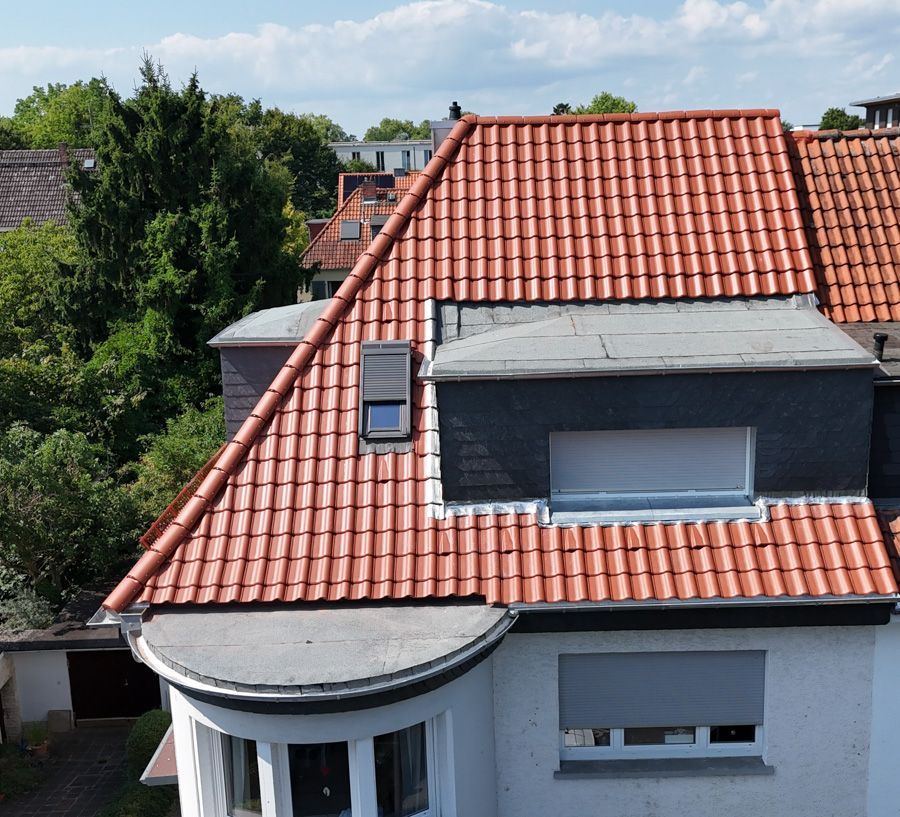 Weißes Haus mit rotem Ziegeldach, kleinem Dachgaubenfenster und geschwungenem Dachteil. Im Hintergrund grüne Bäume.