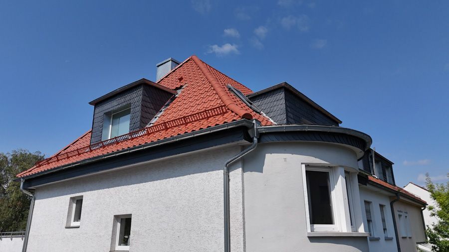 Weißes Gebäude mit rotem Ziegeldach, Dachgauben und grauen Zierelementen vor blauem Himmel.