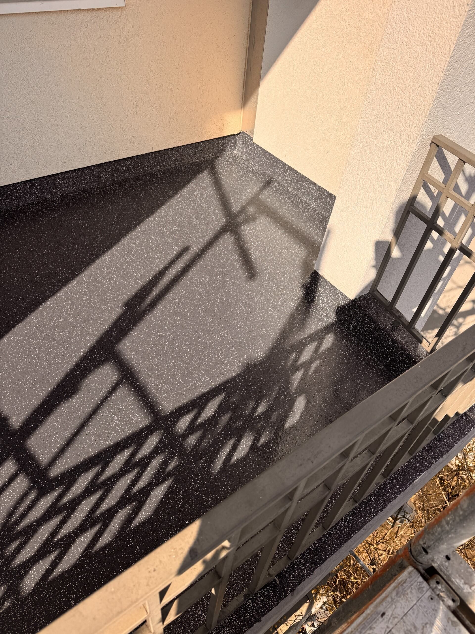 Sonnendurchflutete Dachterrasse mit schwarzem Boden und Metallgeländer, die Schatten auf beige Wände werfen