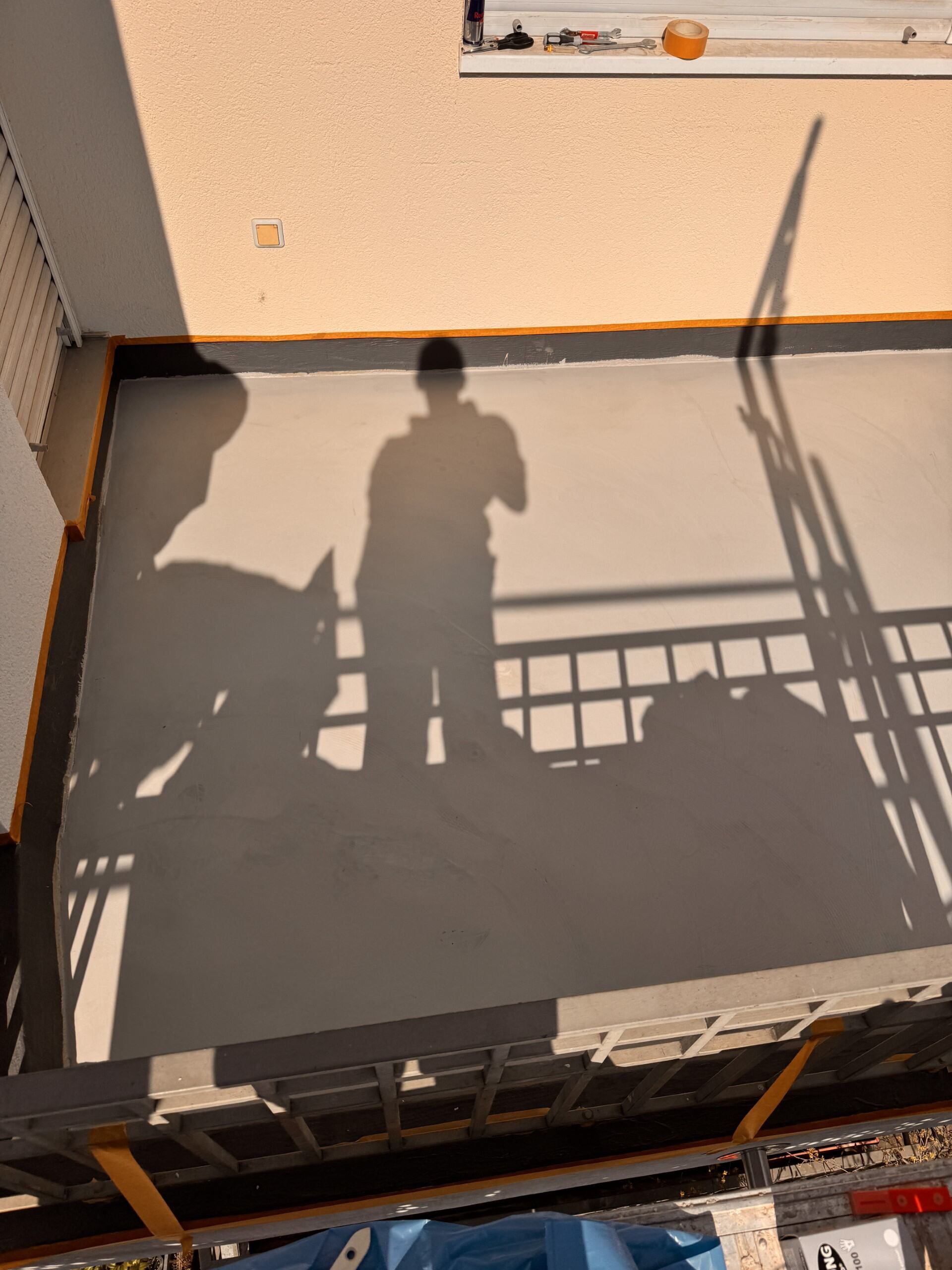 Schatten zweier Personen auf einem sonnenbeschienenen Balkon mit Geländer und beiger Wand