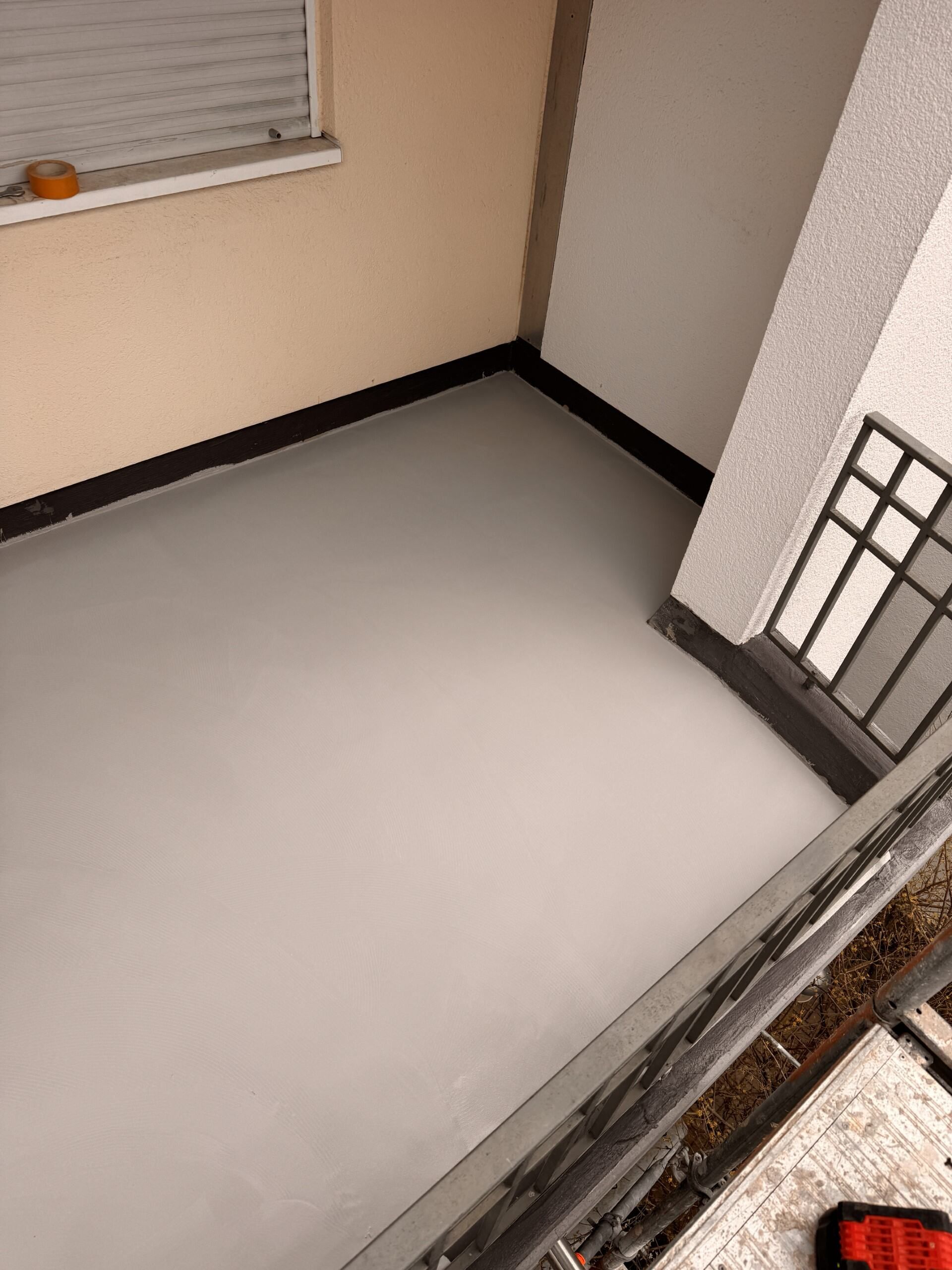Leeres beigefarbenes Eckzimmer mit hellgrauem Boden und dunklen Zierleisten in der Nähe eines Fensters und eines Türgeländers.