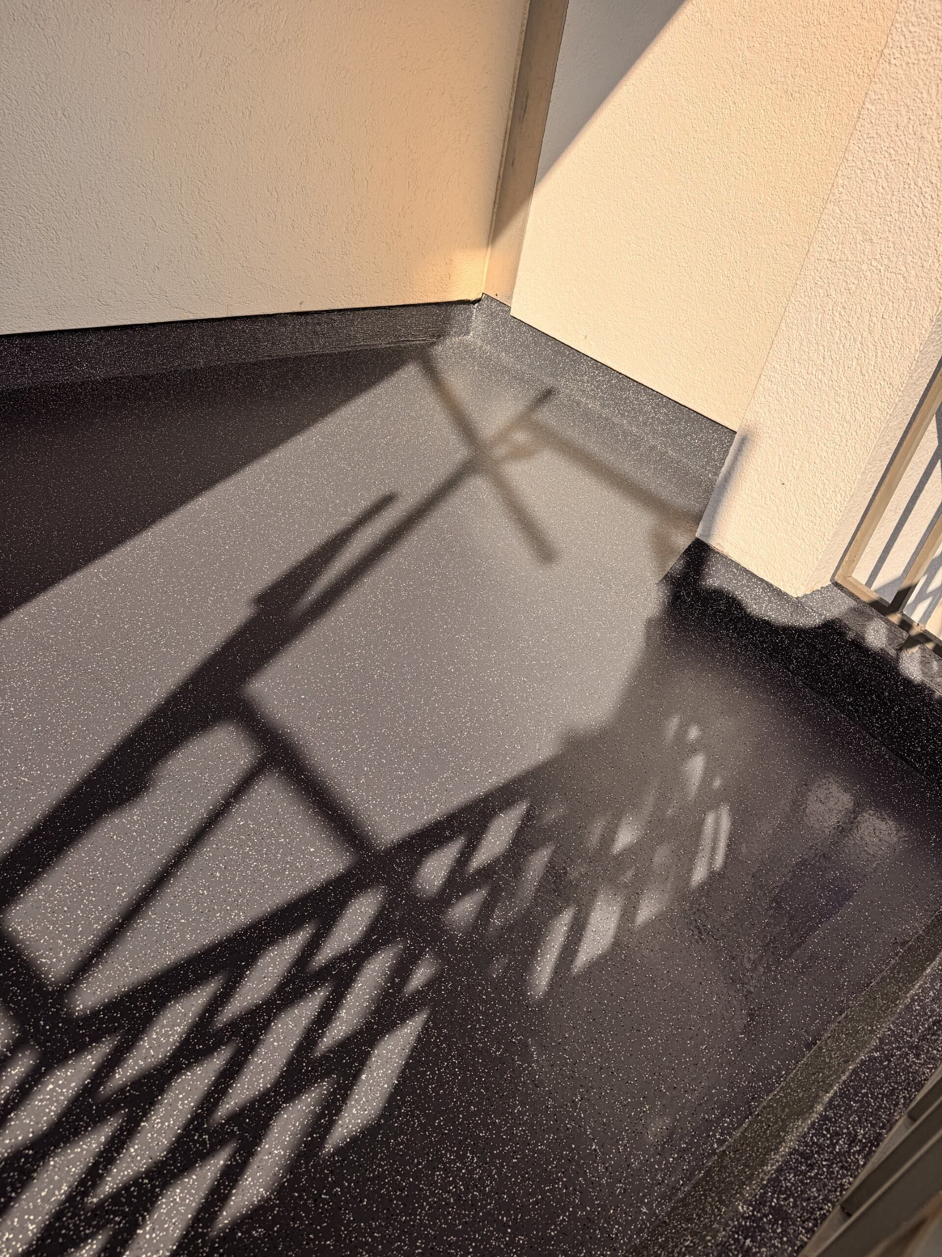 Sonnendurchflutete Balkonecke mit schwarzem Boden und dekorativem Geländer, dessen Schatten auf beige Wände fallen.