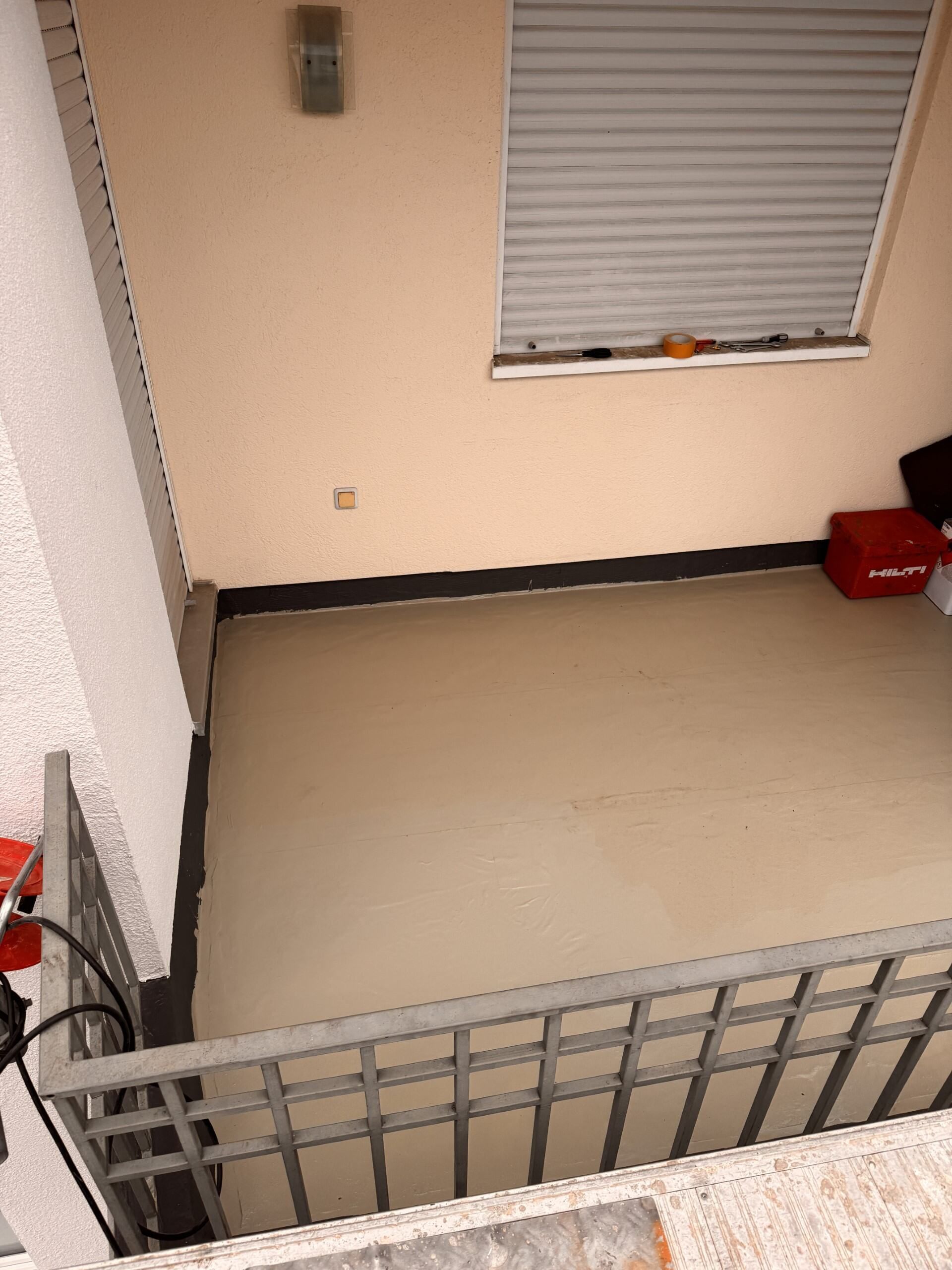 Leerer beigefarbener Raum mit geschlossenem Fensterrollo und grauem Metallgeländer im Vordergrund.