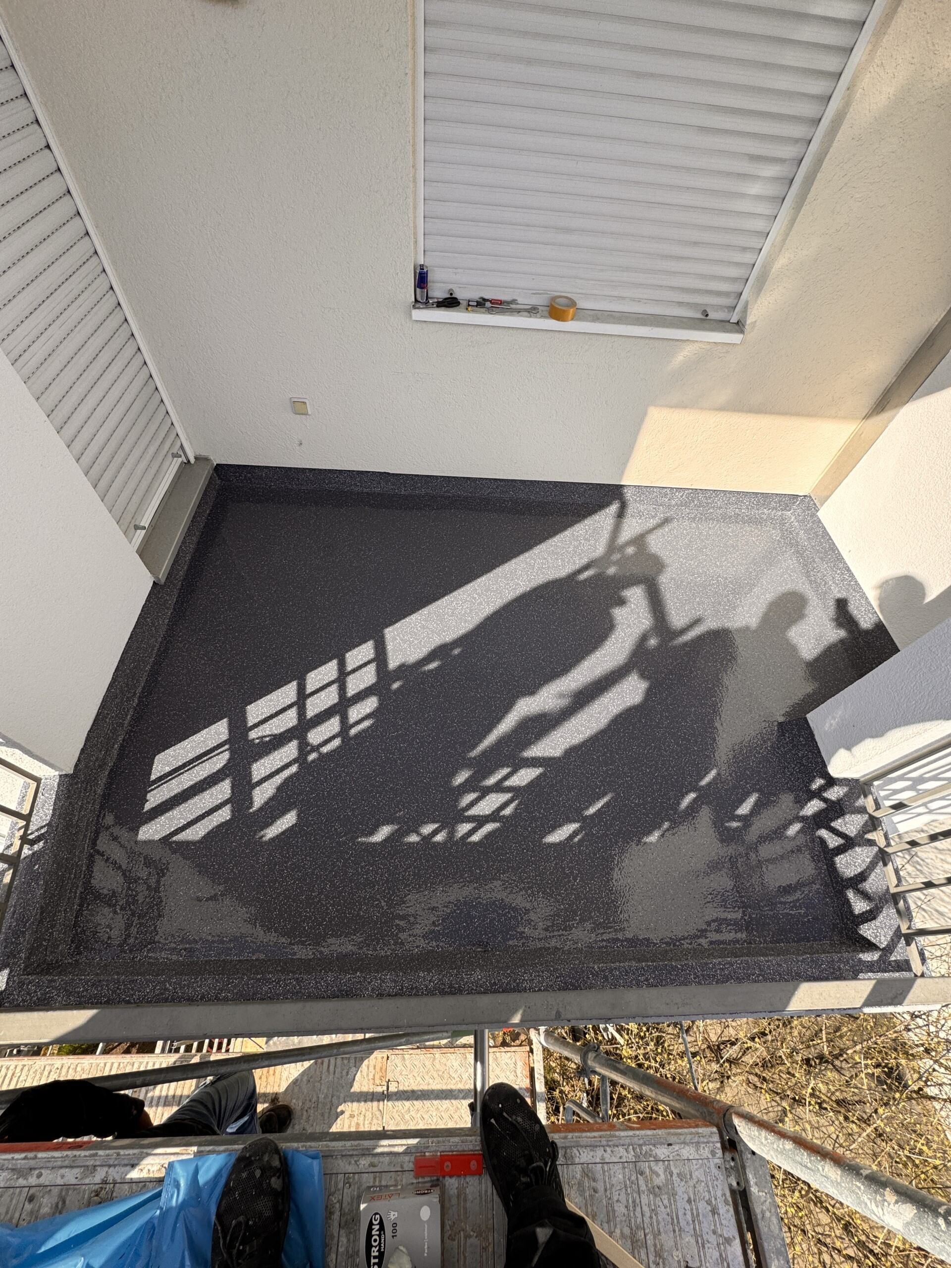 Kleiner Balkon mit dunklem Boden, weißen Wänden und starken Schattenwurf durch die Fensterrahmen im Sonnenlicht.