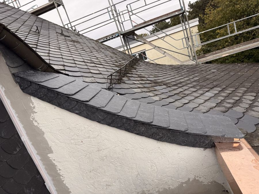 Gebogenes Schieferdach wird renoviert, Gerüst und darunter befindet sich eine weiße Außenwand.