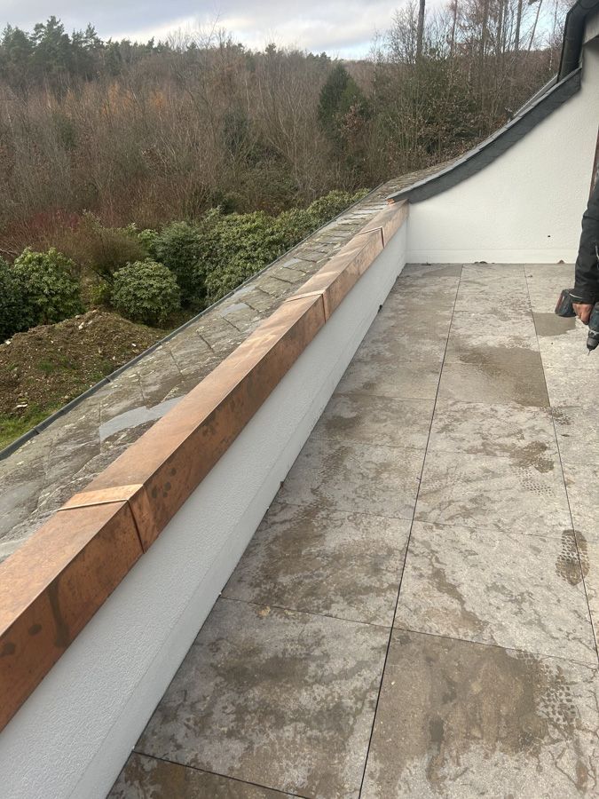 Schmale Dachterrasse mit Steinplatten, Holzgeländer und Blick auf einen bewaldeten Hang.