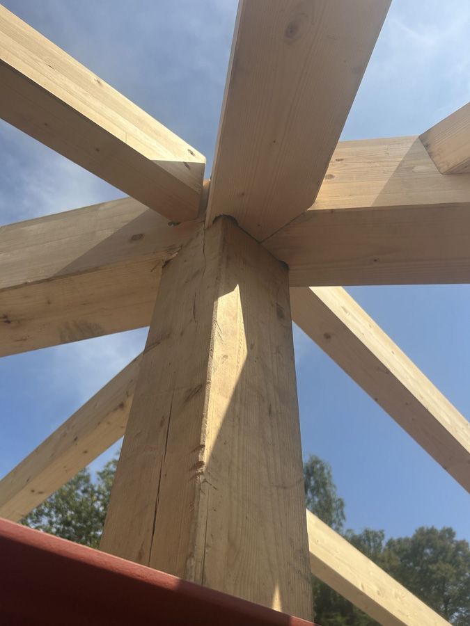 Nahaufnahme einer Holzbalkenverbindung unter blauem Himmel, mit schräg verlaufenden Stützbalken, die von einem zentralen Pfosten ausgehen.