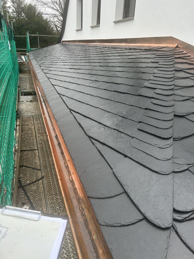 Ein im Bau befindliches Schieferdach befindet sich neben einer weißen Mauer und einem grünen Zaun; am Rand steht ein Gerüst.