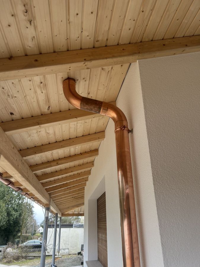 Kupfernes Fallrohr und Dachrinne auf einem hölzernen Verandadach, mit weißer Wand und Blick nach draußen darunter