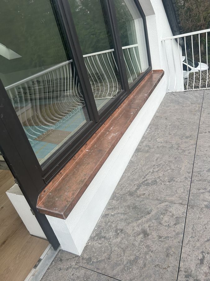 Moderne Gebäudefassade mit dunkel gerahmtem Fenster, Holzfensterbank und gefliester Terrasse neben einem Geländer