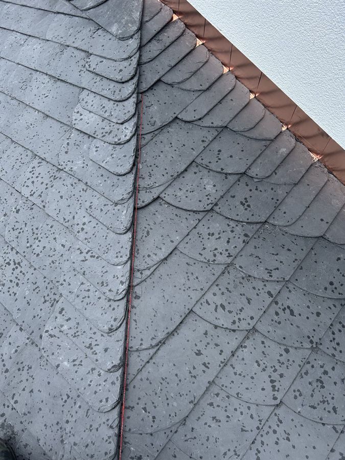 Nahaufnahme eines grauen Schindeldaches mit vertikaler Naht und roter Zierleiste entlang des Dachfirsts.