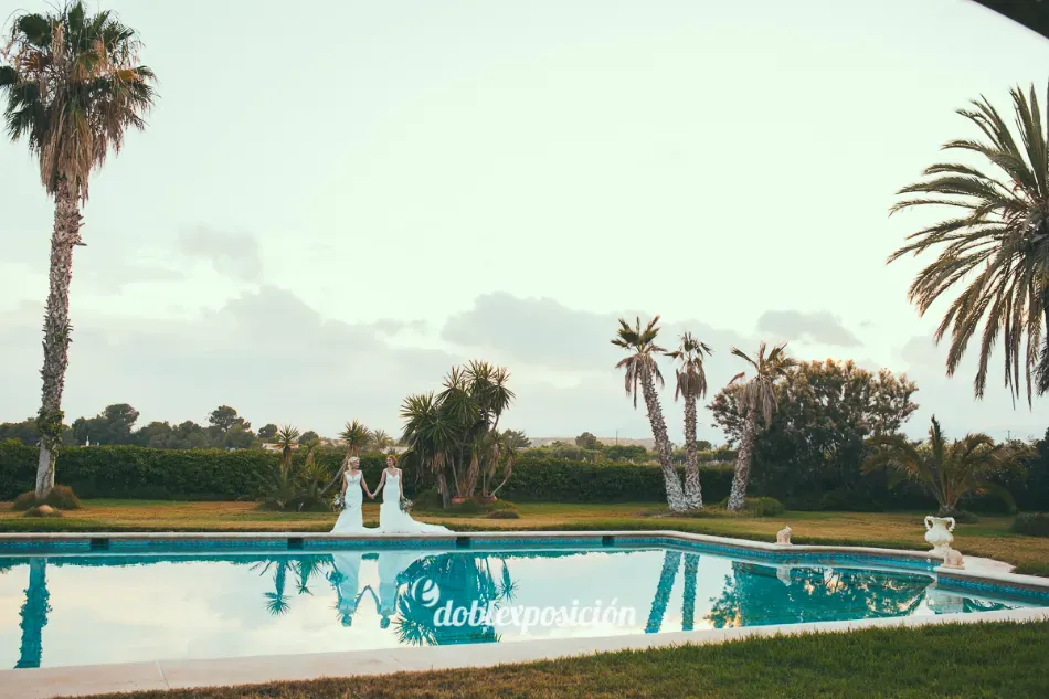 Jardín junto a la piscina con dos personas vestidas de blanco, palmeras y una piscina turquesa al atardecer.