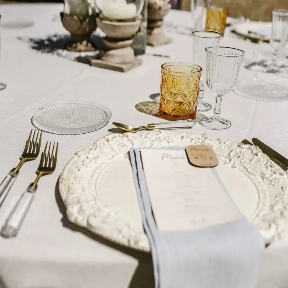 Elegante mesa puesta con platos blancos, cubiertos de plata y copas de color ámbar sobre un mantel blanco.