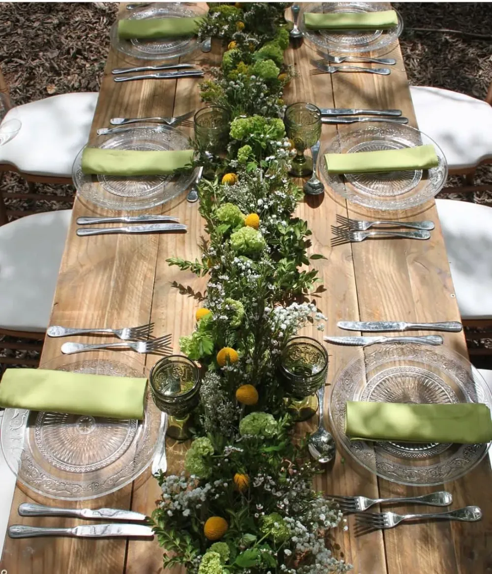 Mesa rústica al aire libre con manteles individuales tejidos, servilletas verdes dobladas y un centro de mesa floral con pequeñas frutas amarillas.