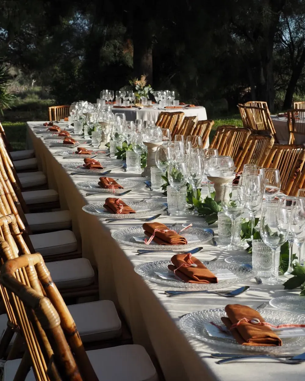 Conjunto de mesa de comedor larga para exteriores con mantelería blanca, cristalería y servilletas marrones en un entorno ajardinado.