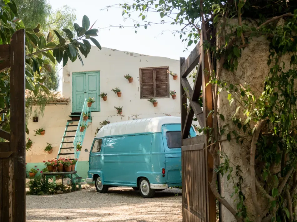 Una furgoneta turquesa aparcada en un patio frondoso junto a una casa blanca con contraventanas de color verde azulado y una escalera.