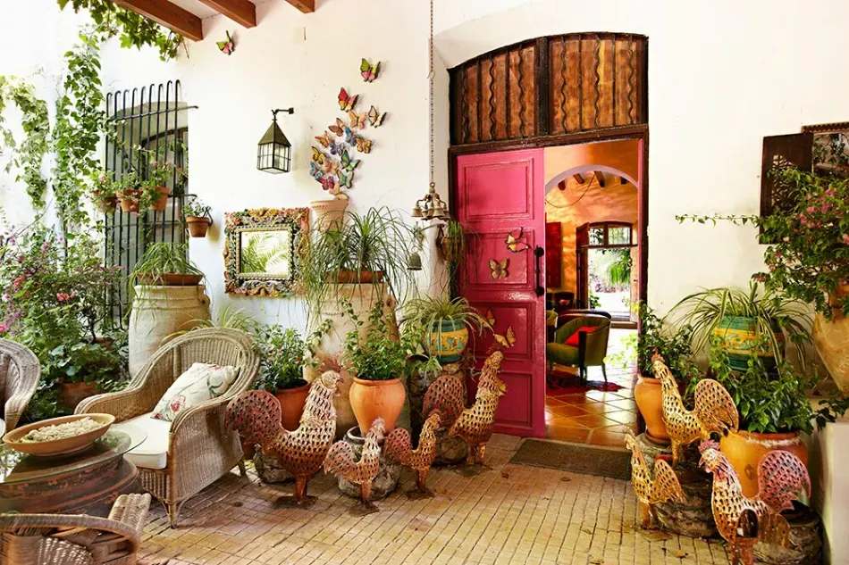 Patio ajardinado colorido con plantas en macetas, sillas de mimbre y una puerta arqueada de color rosa.
